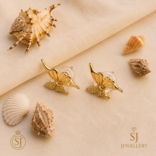 Gold-tone butterfly stud earrings with crystal-studded body, displayed on cream fabric with seashells
