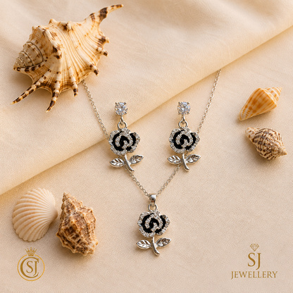 Silver-tone black rose pendant necklace and matching drop earrings with crystal stone detailing, displayed on cream fabric with seashells